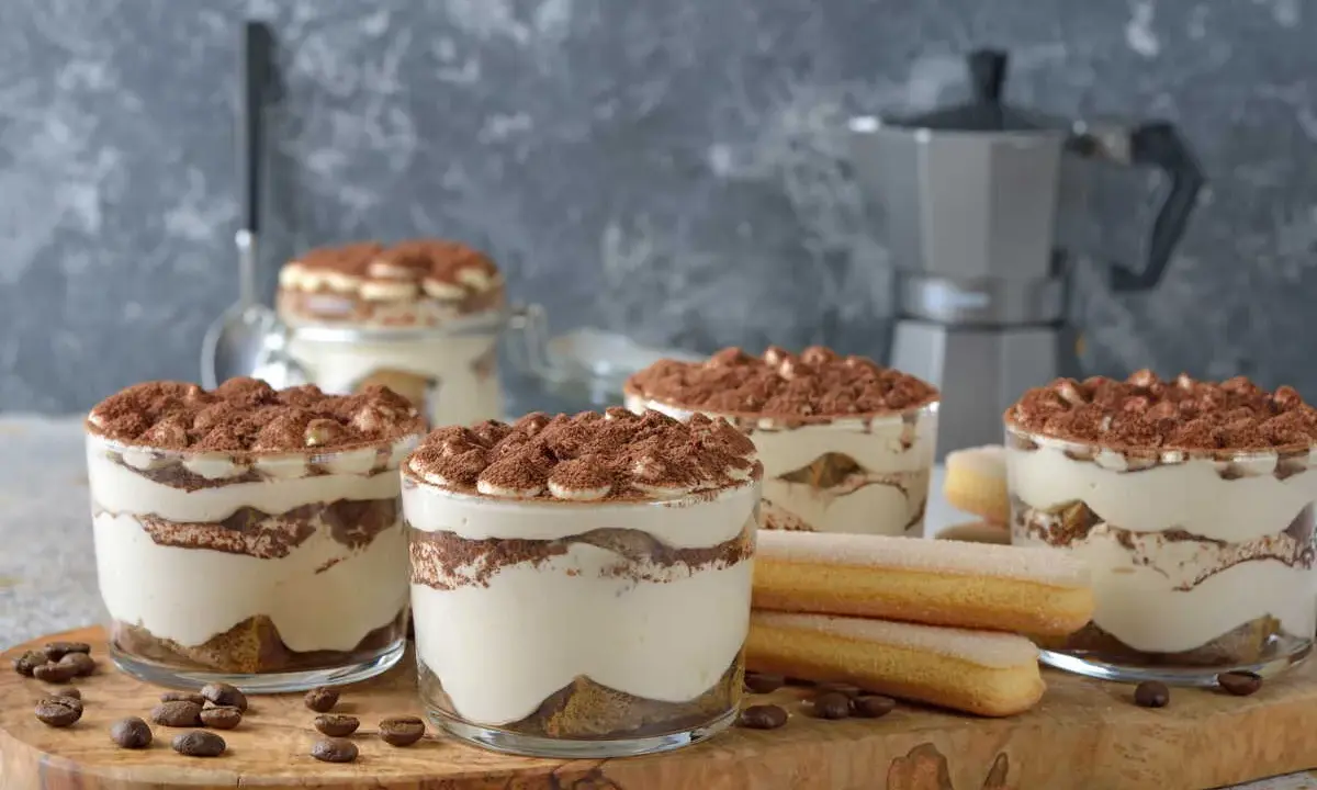 Plusieurs verrines de tiramisu sans café, garnies de cacao, côtoient des biscuits à la cuillère et des grains de café sur une planche en bois.