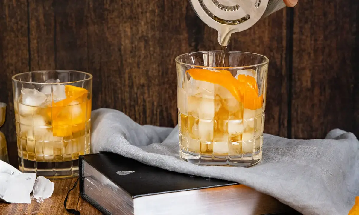 Ein Old Fashioned wird zubereitet, Eiswürfel und Orangenschale zieren das Glas. Ein altes Rezept wird lebendig.