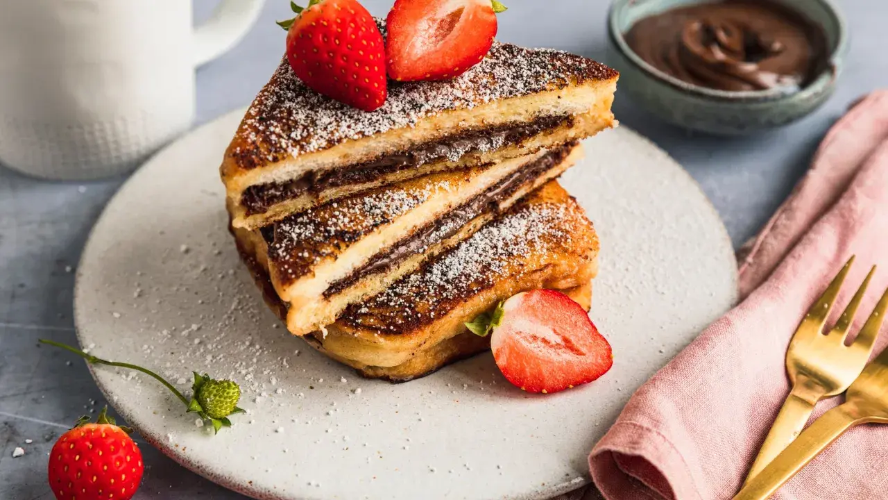 Gestapelte French Toast mit Nutella, bestäubt mit Puderzucker und garniert mit frischen Erdbeeren. Ein Glas Milch und eine Schüssel Nutella im Hintergrund.