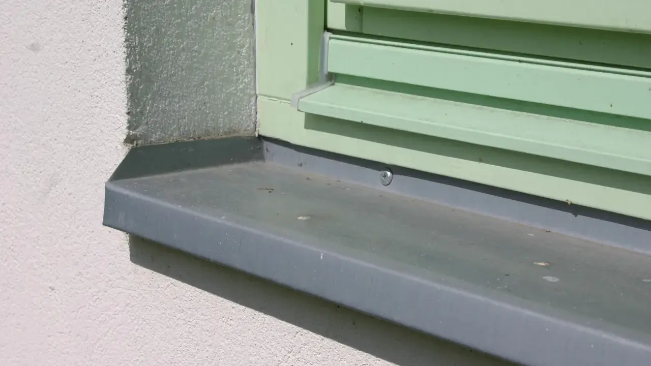 Szary parapet zewnętrzny zamontowany przy oknie z zielonymi okiennicami. Idealny moment, kiedy montować parapety zewnętrzne, to po ociepleniu elewacji.