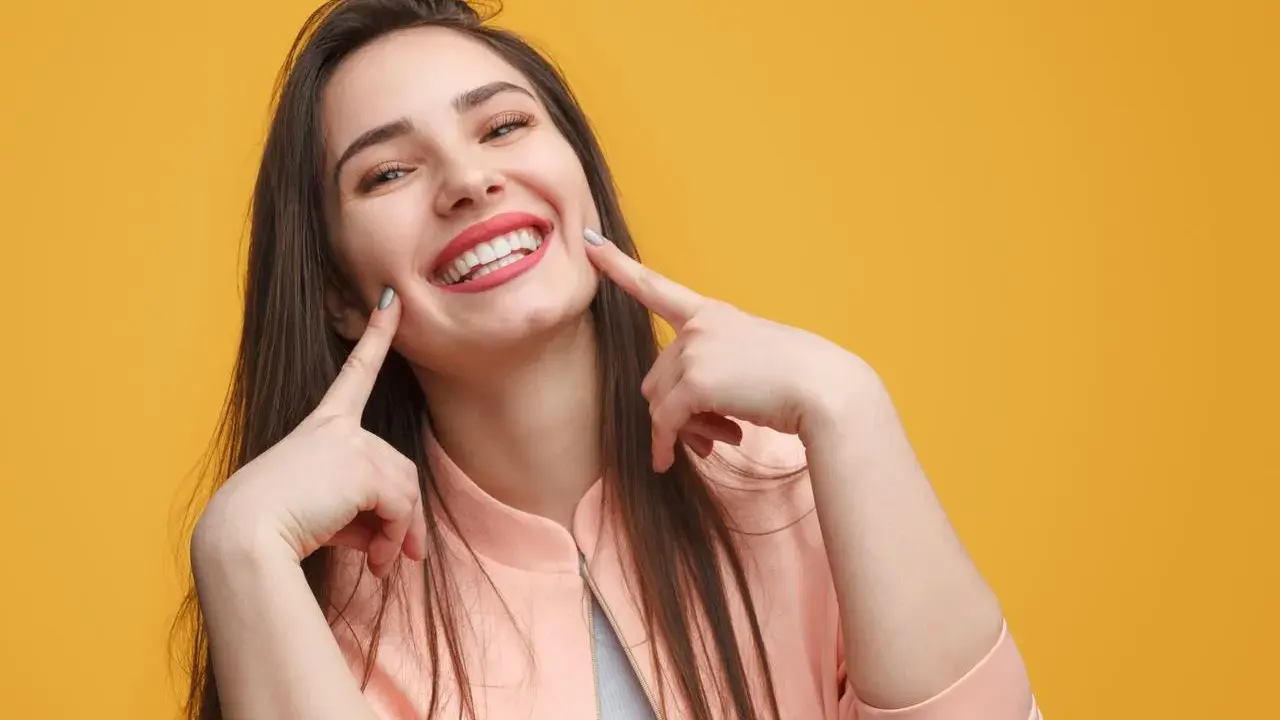 Une jeune femme au sourire éclatant, doigts pointés vers ses joues, semble expliquer la signification d'un sourire pincé.