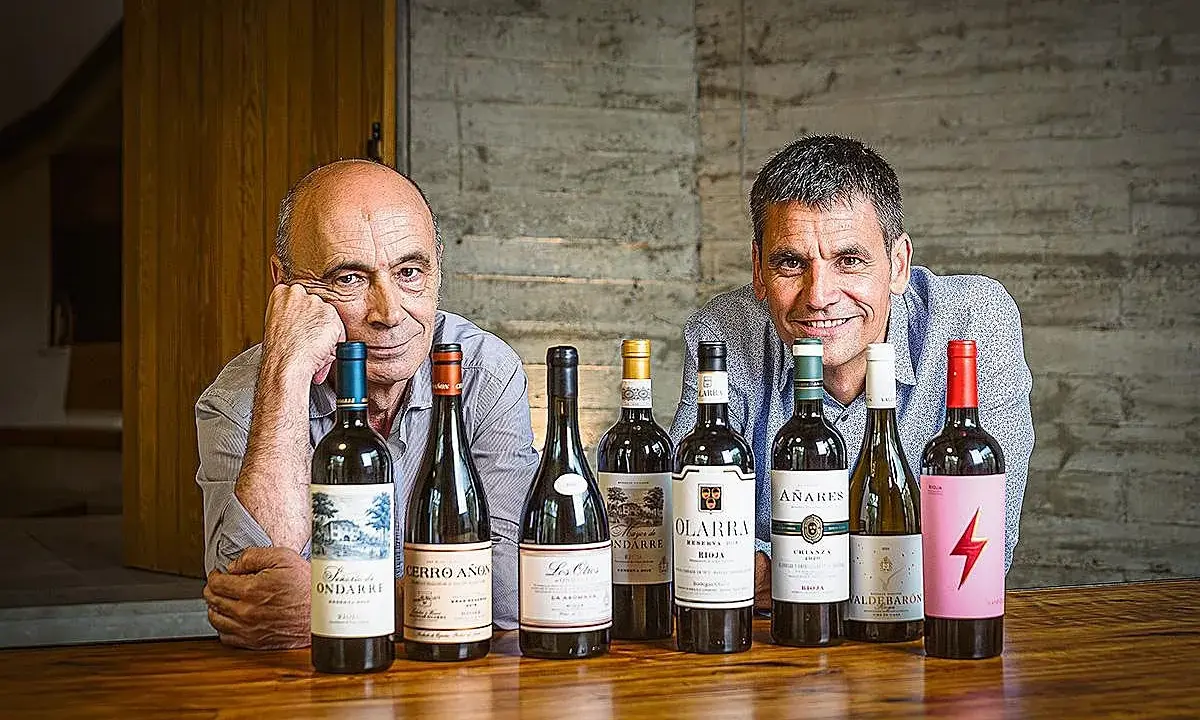 Dos hombres sonríen detrás de una fila de botellas de vino. Uno de ellos es el dueño de Bodegas Olarra.