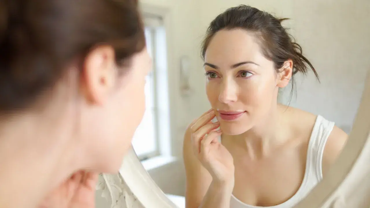 Une femme examine son visage dans un miroir, touchant sa joue. Sa peau semble très fine, peut-être signe d'une carence.