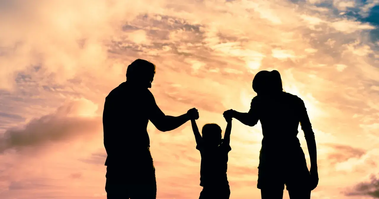 Siluetas de una familia jugando bajo un cielo anaranjado. Un niño es levantado por sus padres, un ejemplo de aprendizaje social y emocional.