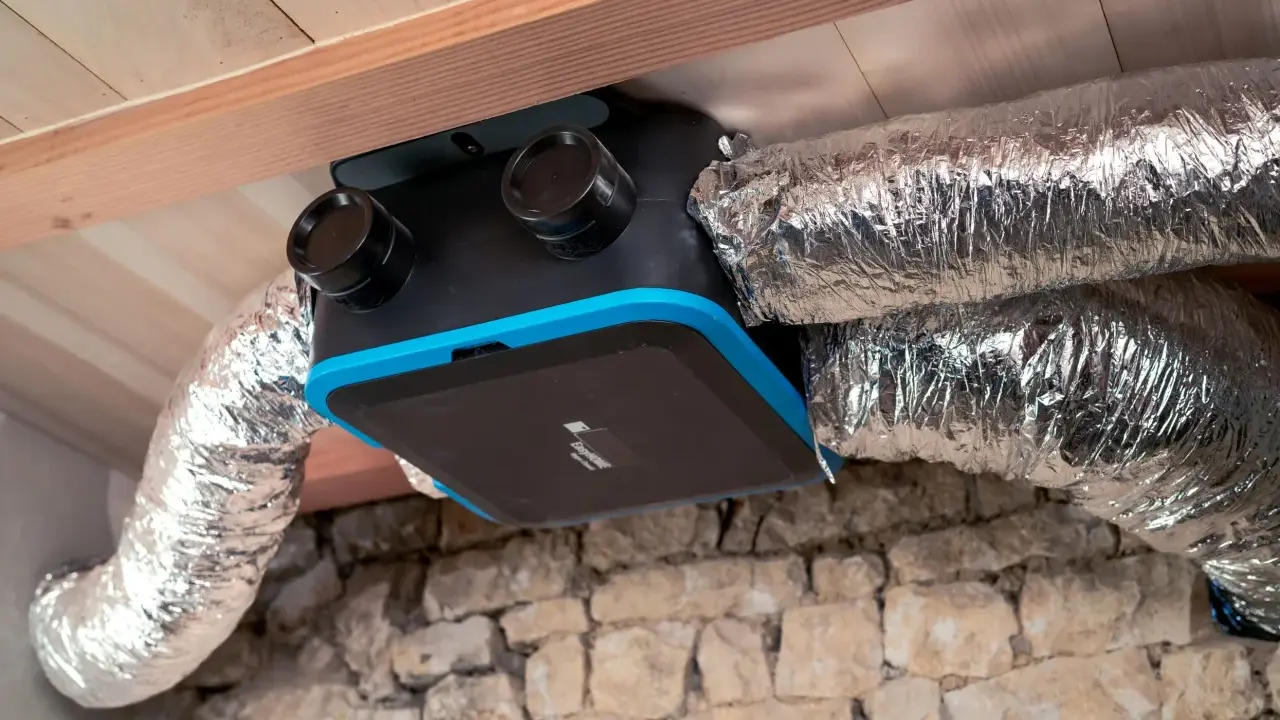 Unité de ventilation maison avec conduits argentés, installée sous un plafond en bois et au-dessus d'un mur en pierre.