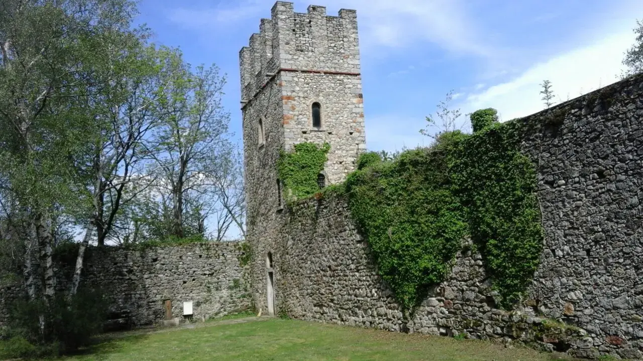 Antica torre del forte di Orino, circondata da mura in pietra e vegetazione rigogliosa, sotto un cielo azzurro.
