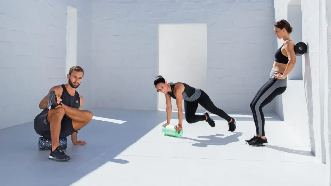 Drei Sportler nutzen eine **blackroll vor oder nach dem Training**. Ein Mann dehnt sich, eine Frau macht Mountain Climbers, die andere lehnt sich an die Wand.