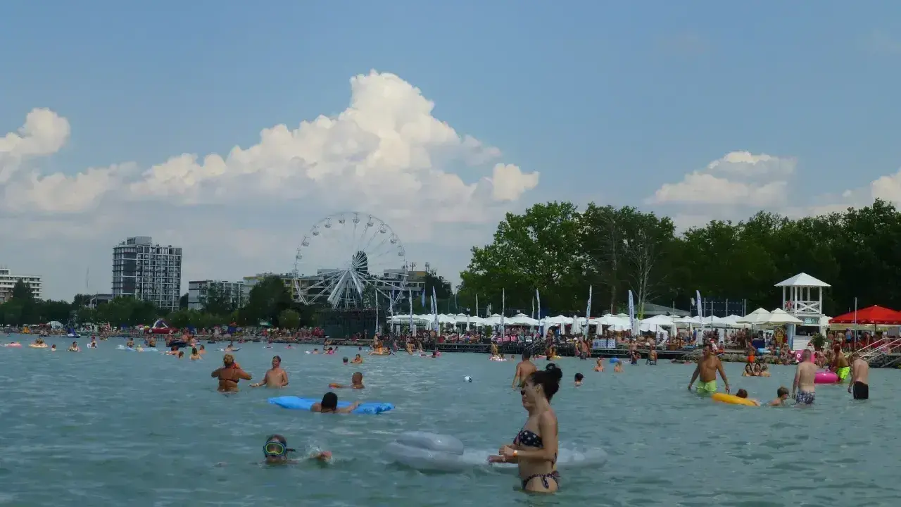 Tłum ludzi cieszy się słońcem i wodą na **Balaton plaża**. W tle widać diabelski młyn i budynki.