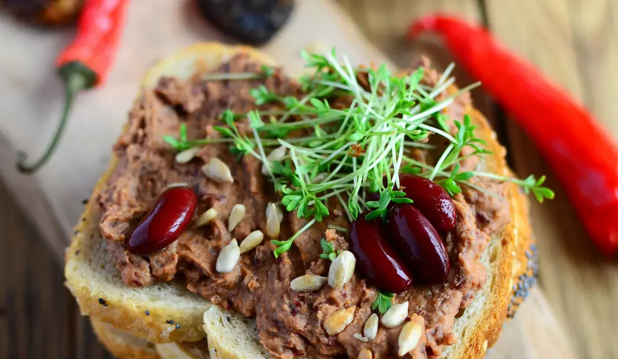 Pyszna pasta z czerwonej fasoli na chlebie, ozdobiona kiełkami i nasionami. Obok leżą czerwone papryczki chili.