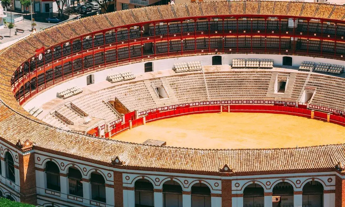 Leere Plaza de Toros mit roten Geländern und sandiger Arena. Die Sitzreihen sind leer, die Sonne scheint auf die traditionelle Architektur.