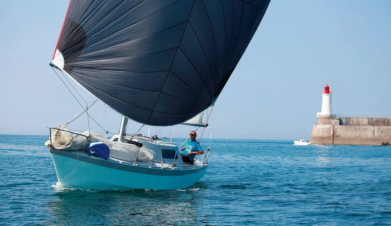 Un voilier bleu avec une grande voile noire navigue près d'un phare. Le skipper, portant un t-shirt bleu, semble observer un ovni 25 dans le ciel clair.