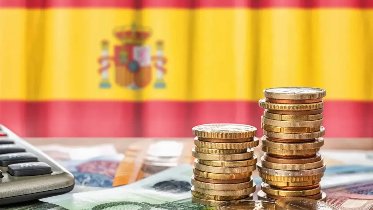 Monedas y billetes sobre una mesa, con la bandera de España de fondo. Una calculadora a la izquierda. Reflexión sobre qué son los tributos.