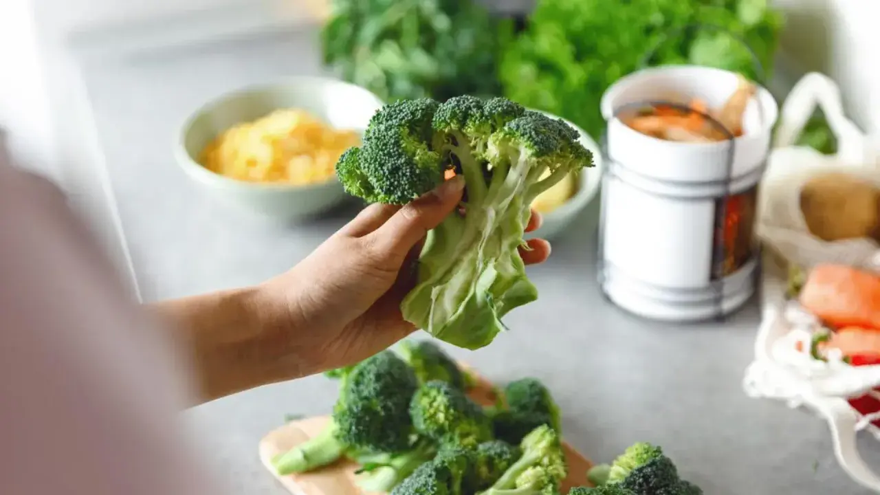 Mano sosteniendo un brócoli fresco, listo para preparar deliciosas recetas de brócoli. En el fondo, queso rallado y zanahorias.