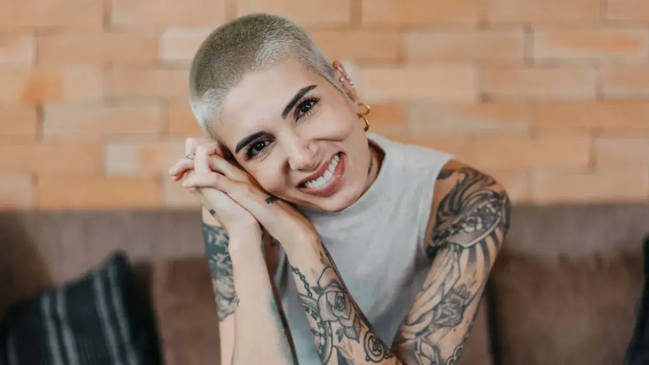 Lächelnde Frau mit Buzz Cut und vielen Tattoos, die ihre Hände vor dem Gesicht verschränkt.