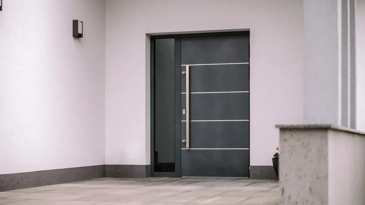 Nowoczesne drzwi wejściowe, ciemnoszare, z długą, metalową klamką i bocznym przeszkleniem. Czy to drzwi stalowe czy aluminiowe?