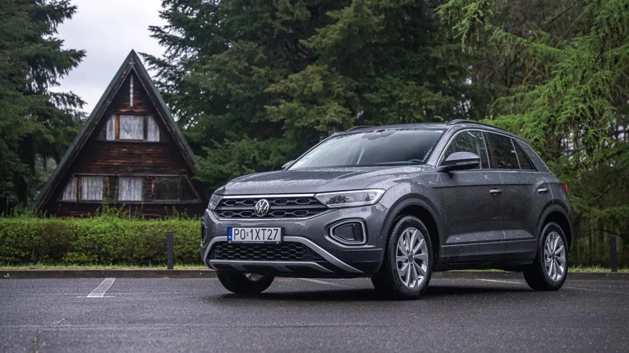 Szary Volkswagen T-Roc używany, zaparkowany przed drewnianym domkiem w lesie. Idealny na wypady za miasto.