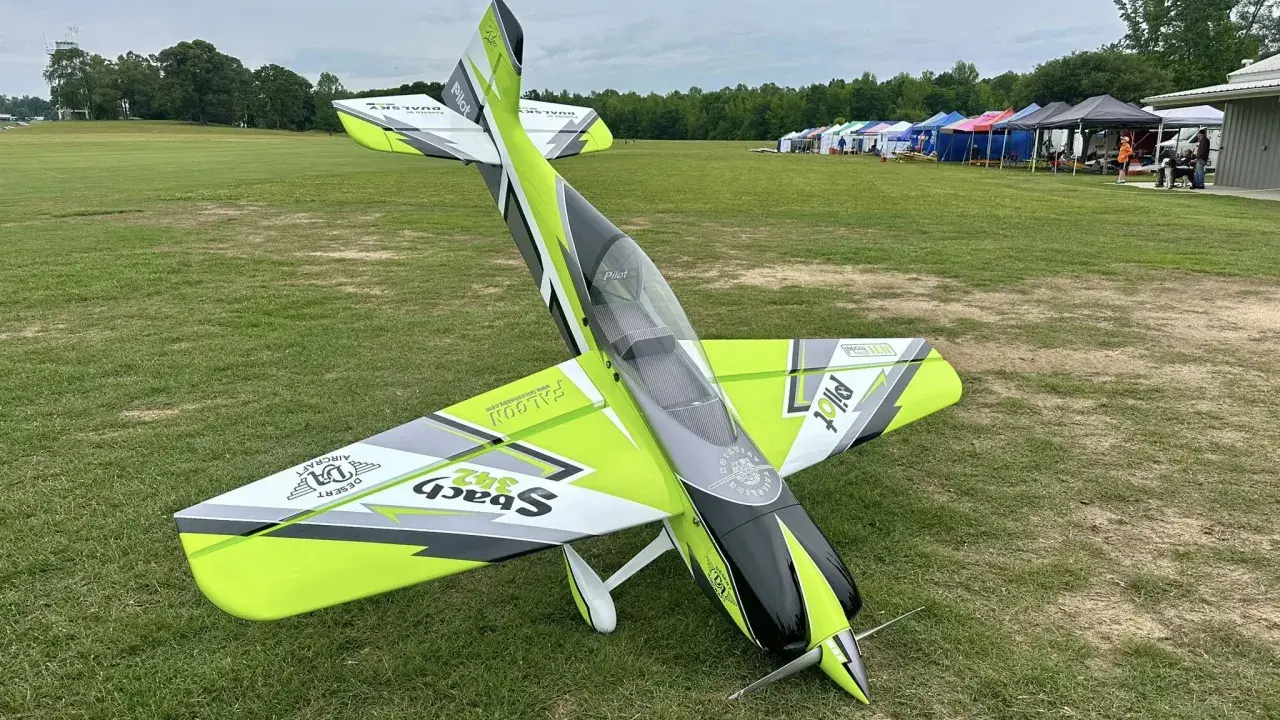 Ein ramy rc-Flugzeug mit auffälligem Design steht auf einer grünen Wiese. Im Hintergrund Zelte und Bäume.