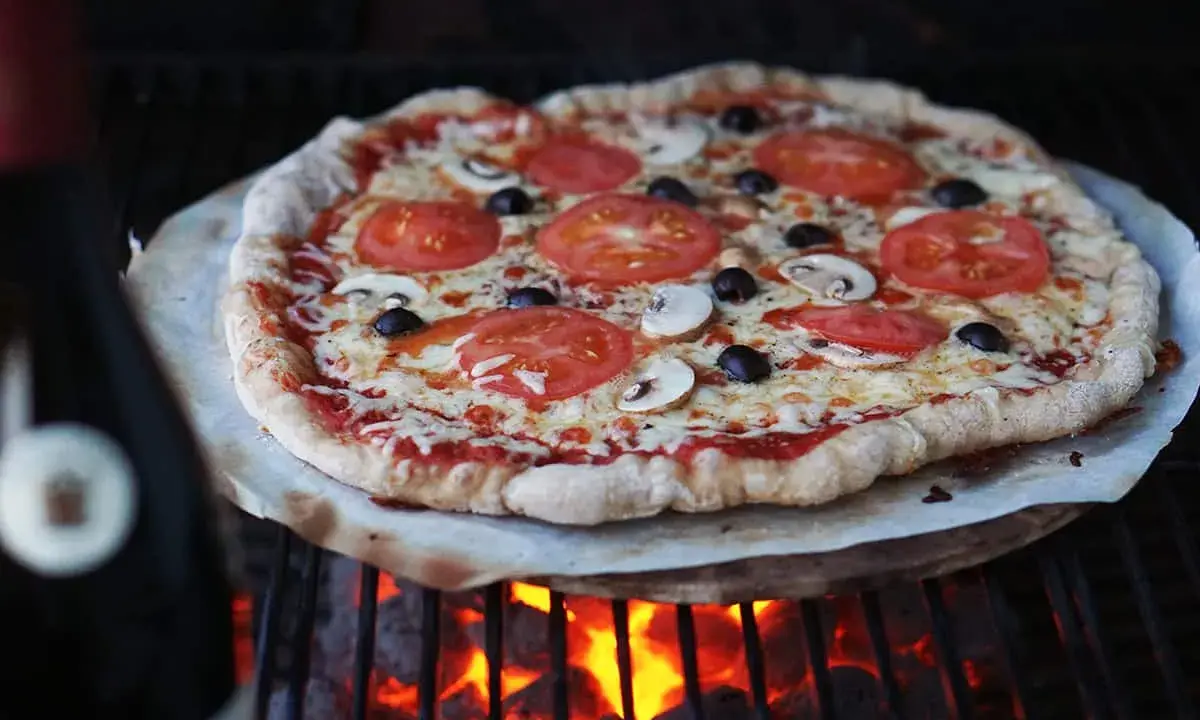 En hemmagjord pizza med tomater, champinjoner och oliver grillas över glödande kol. Perfekt för ett italiensk pizzadeg recept.