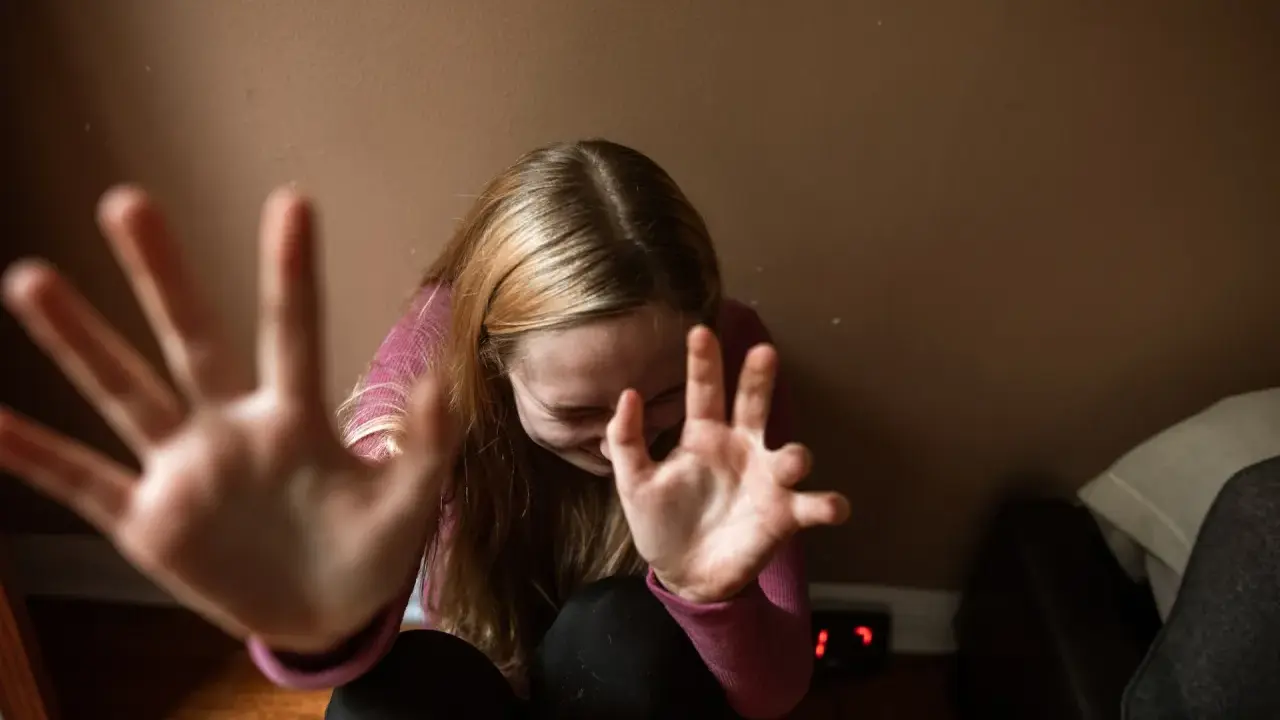 Dziewczyna z rozczochranymi włosami, z rękami przed twarzą, jakby broniła się przed nadmierną kontrolą.