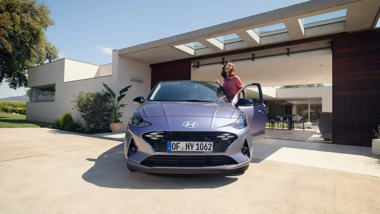 Mujer sonriente junto a un coche alto y morado, aparcado frente a una casa moderna con puertas abiertas.