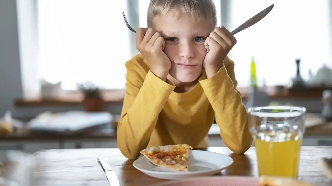 Chłopiec z widelcem i nożem w dłoniach, z miną mówiącą "dziecko nie chce jeść". Przed nim kawałek pizzy i szklanka soku.