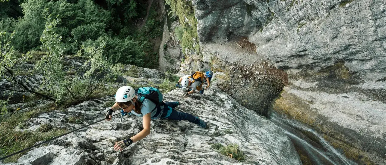 Osoby w kaskach i uprzężach pokonują skalną ścianę, używając liny i karabinków. Cały niezbędny via ferrata sprzęt zapewnia im bezpieczeństwo podczas wspinaczki.