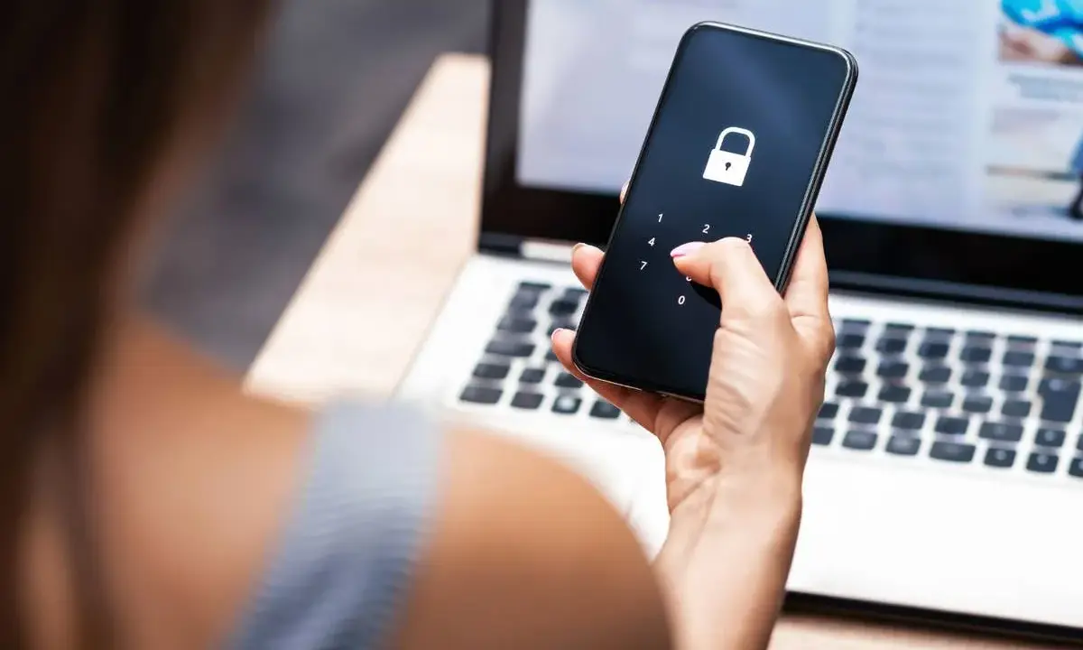 Frau entsperrt Handy mit Schloss-Symbol, besorgt wegen gehackter mailadresse. Laptop im Hintergrund.