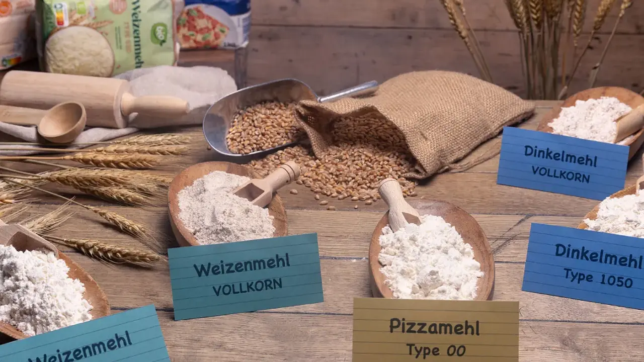 Verschiedene Mehlsorten, darunter Weizenmehl, Dinkelmehl und Pizzamehl, liegen in Holzschalen. Daneben Weizenkörner und ein Nudelholz.