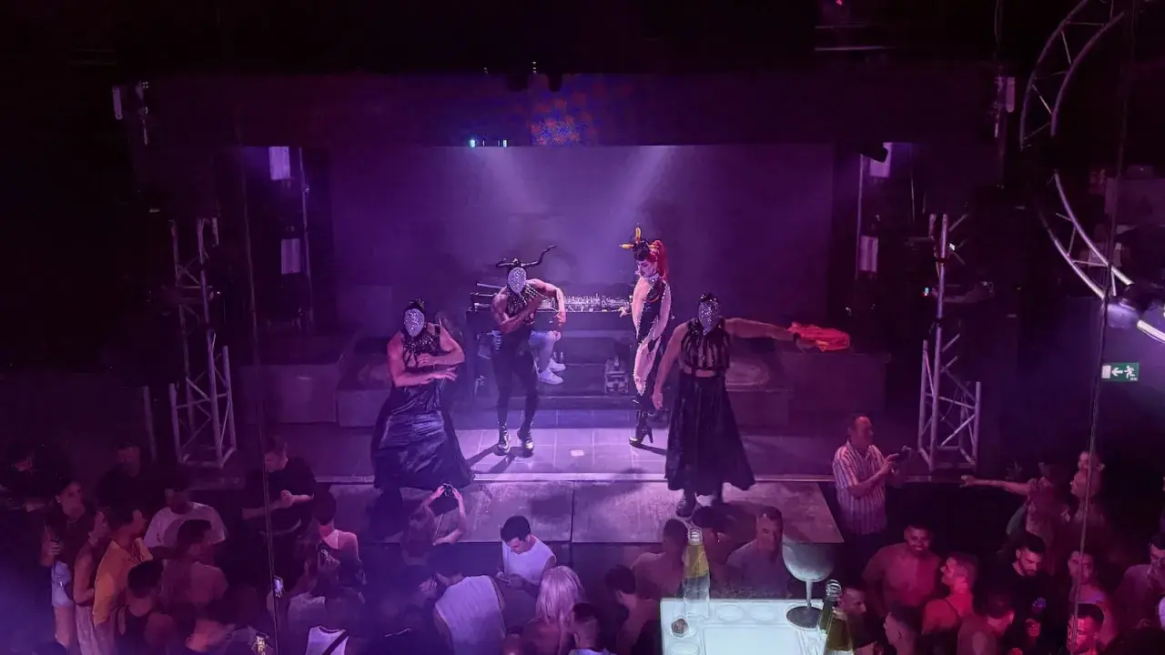 Performers in masks and elaborate outfits dance on stage at one of Valencia's vibrant gay bars, while a crowd watches below.