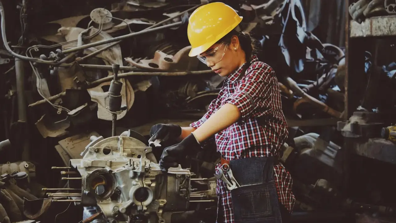 Mujer con casco amarillo y gafas de seguridad trabajando en un motor. Usa guantes negros y un delantal de mezclilla, demostrando el uso de los equipos de protección individual.