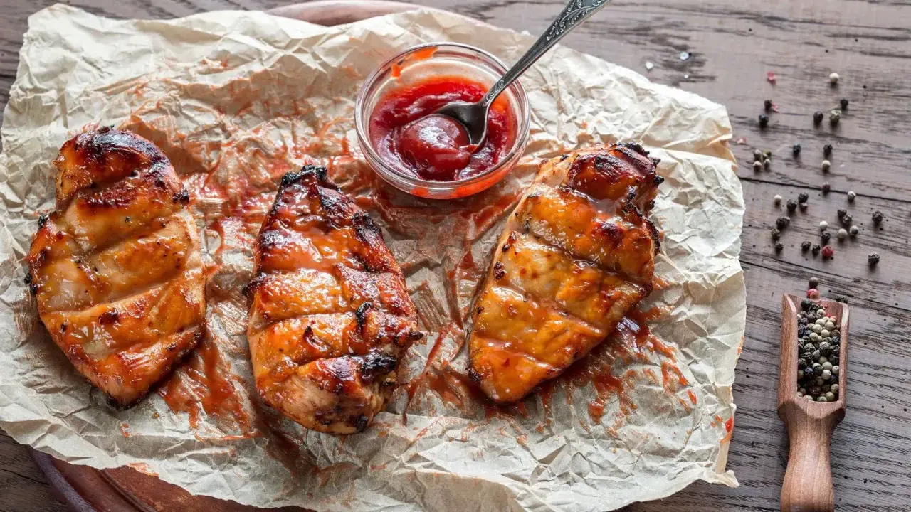 Saftiges Grillfleisch zart machen: Drei gegrillte Hähnchenbrustfilets mit Sauce auf Backpapier, daneben eine Schale Ketchup und eine Holzschaufel mit Pfefferkörnern.