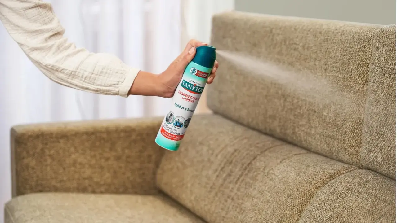 Mano rociando desinfectante en spray sobre un sillón de tela. Una forma fácil de saber como limpiar sillones de tela y eliminar olores.