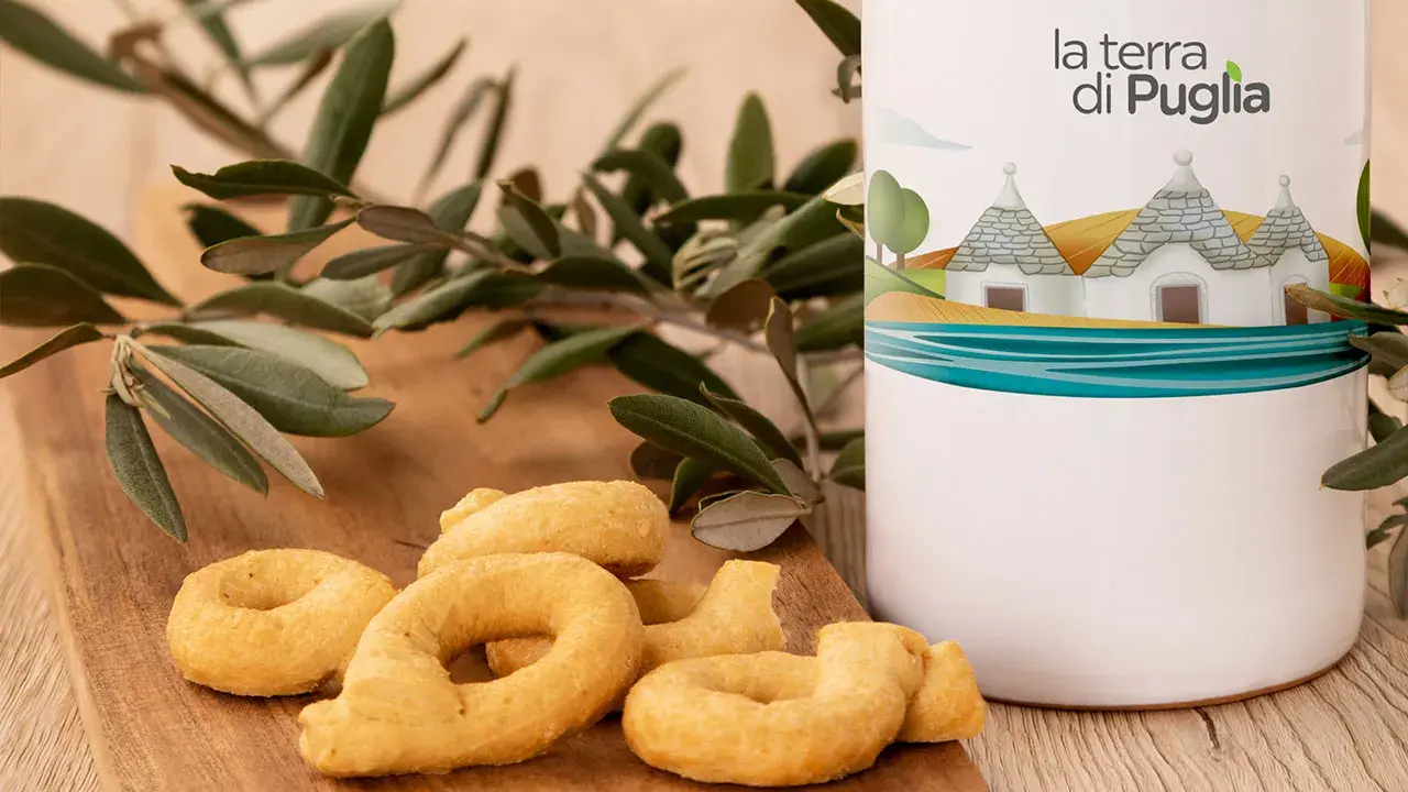 Taralli pugliesi su tagliere di legno, accanto a un contenitore con il logo "la terra di Puglia" e un ramo d'ulivo.