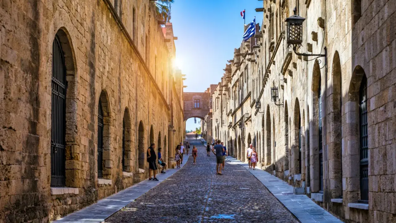 Sonnenbeschienene Gasse in Rhodos Stadt mit Kopfsteinpflaster und historischen Gebäuden. Touristen schlendern durch die mittelalterliche Stadt.