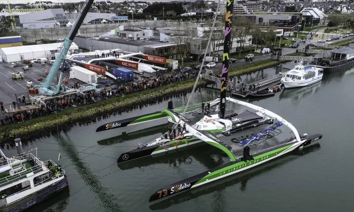 Le trimaran Sodebo Ultim 3, prêt pour la course, est amarré dans un port, entouré de spectateurs et de grues.