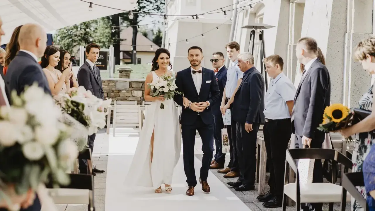 Para Młoda idzie do ołtarza podczas ceremonii ślubnej. Panna Młoda w białej sukni z bukietem kwiatów, Pan Młody w granatowym garniturze.