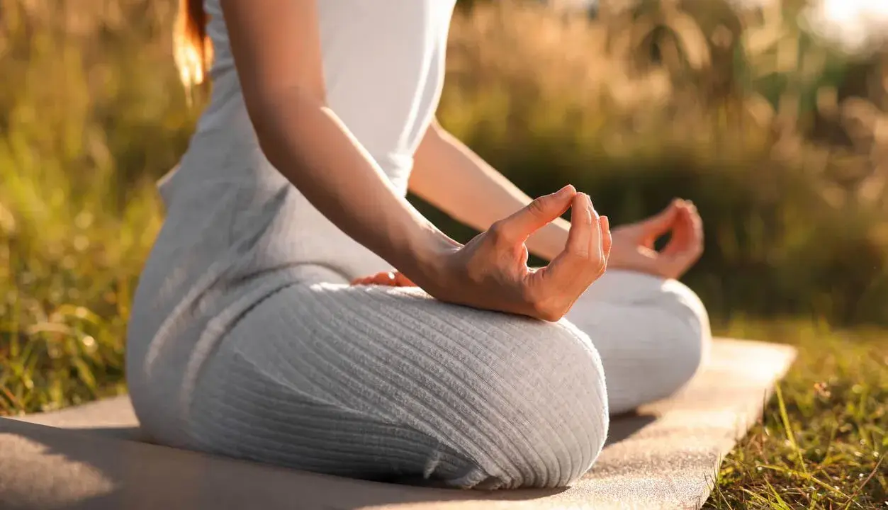 En person sitter i lotusställning på en matta utomhus, med händerna i meditation. Solen lyser genom gräset och skapar en varm atmosfär för morgonmeditation.