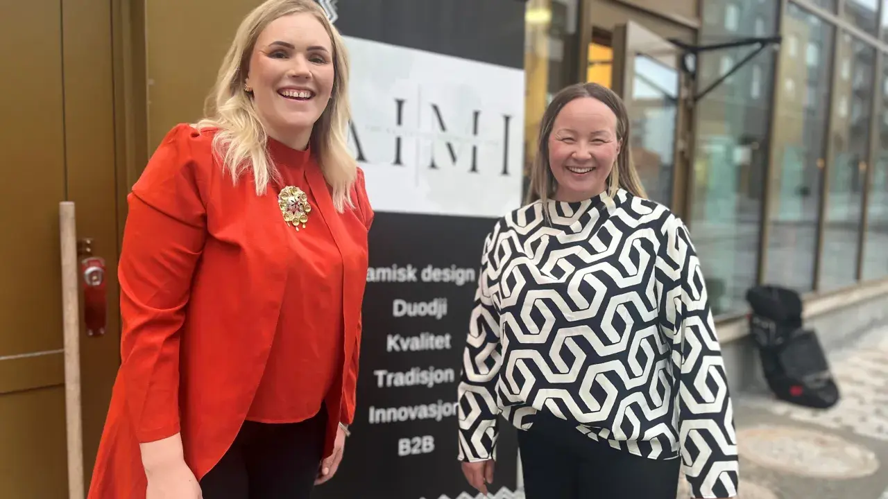 Två glada kvinnor framför en banderoll med text om samisk design och duodji. Kanske säljer de samiska smycken i Kiruna.
