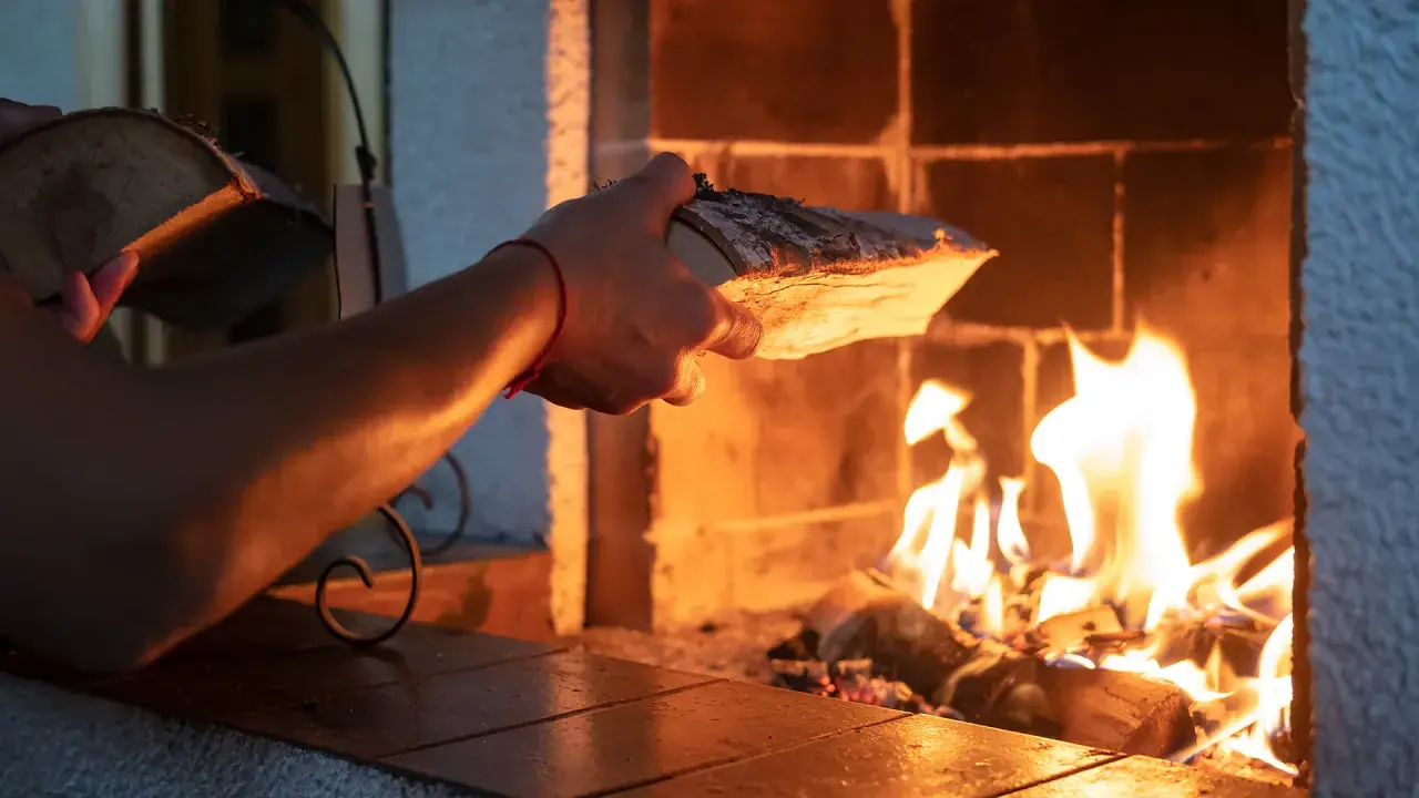 En hand lägger ved i en sprakande brasa. Det syns tecken på soteld i eldstaden.