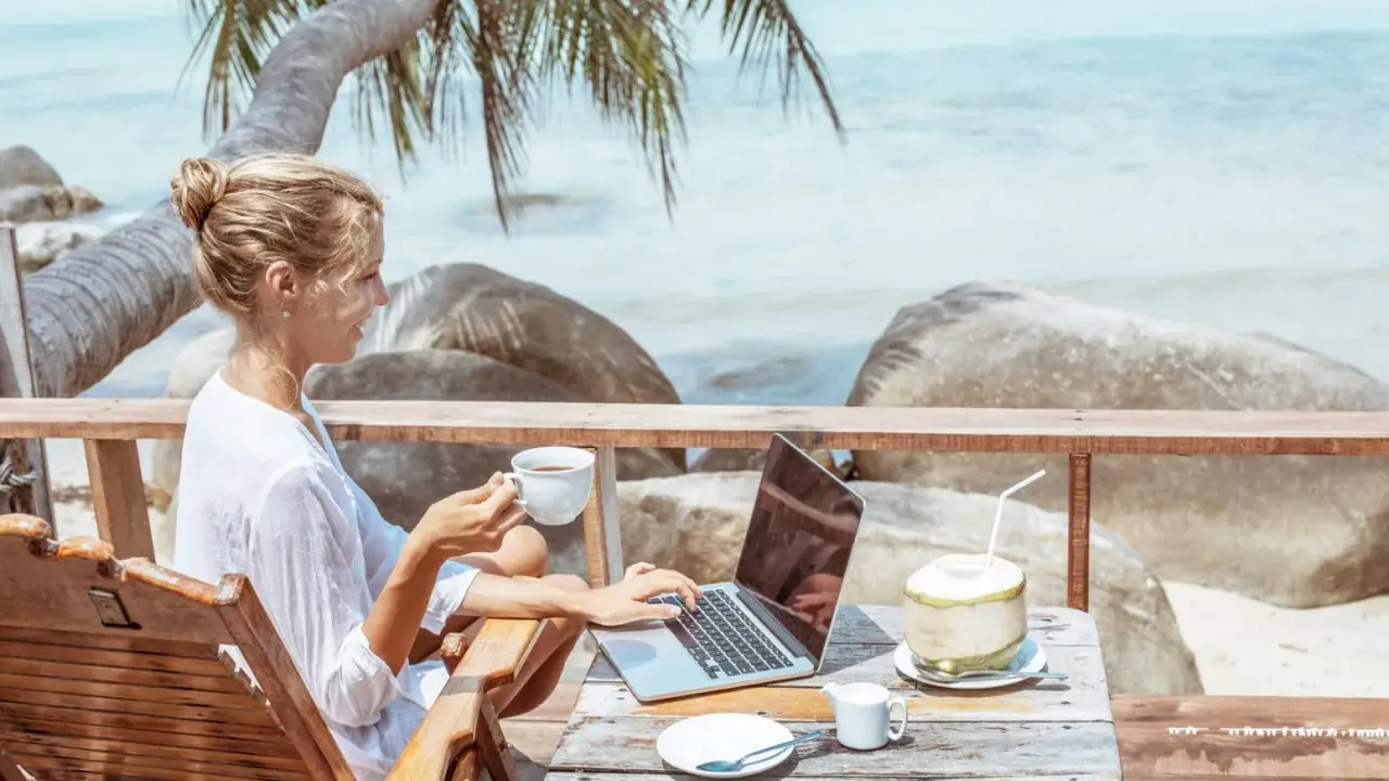 Kvinna med kaffe och laptop på en strandterrass, drömmen om ett digitalt nomad jobb.