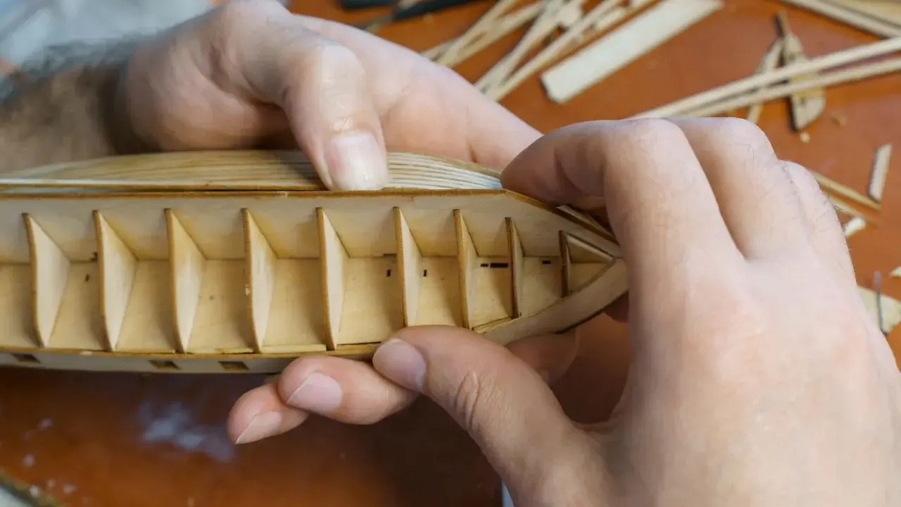 Hände bauen einen hölzernen Schiffsrumpf. Die Rippen des Kaders sind sichtbar, während die äußere Hülle geformt wird.