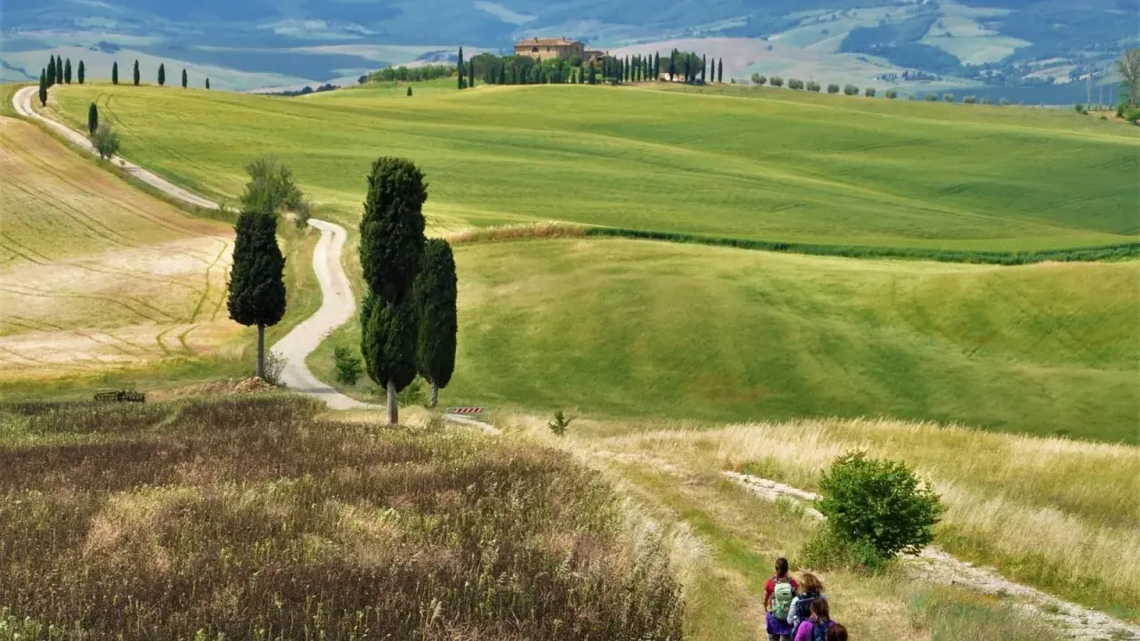 Un gruppo di escursionisti vive le **esperienze Val d'Orcia** su un sentiero tra colline verdi e cipressi, con una casa colonica in lontananza.