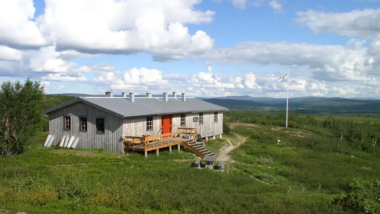 Kungsleden stugor i fjällandskap med molnig himmel. Enkel träbyggnad med veranda och flaggstång.