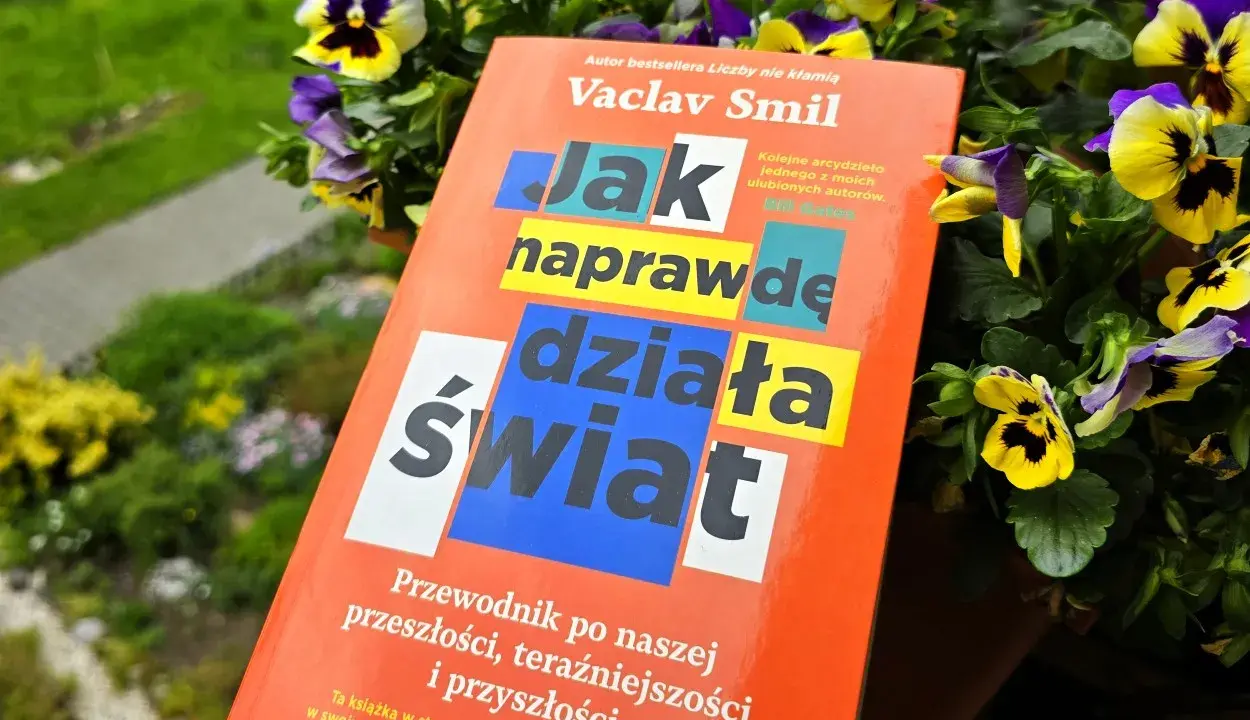 Książka Vaclava Smila "Jak naprawdę działa świat" wśród kwiatów. To przewodnik po przeszłości, teraźniejszości i przyszłości, który pomaga zrozumieć, co to odwyk od błędnych przekonań.