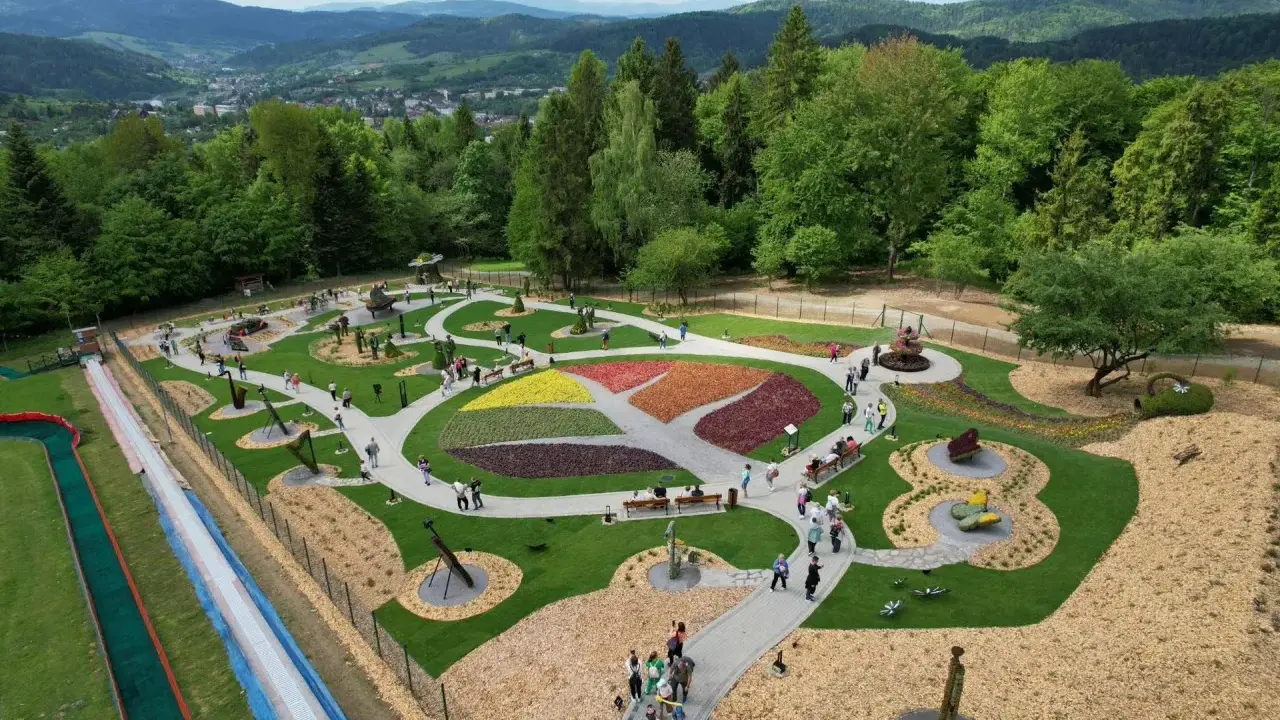 Widok z góry na park z kolorowymi rabatami kwiatowymi i rzeźbami. Widać ścieżki, ławki i ludzi spacerujących po terenie. W tle góry i las.