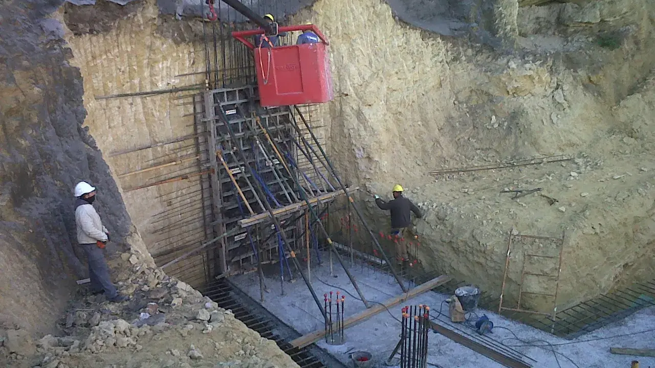 Trabajadores instalan encofrado para muro pantalla, con bataches y estructura de acero en obra.