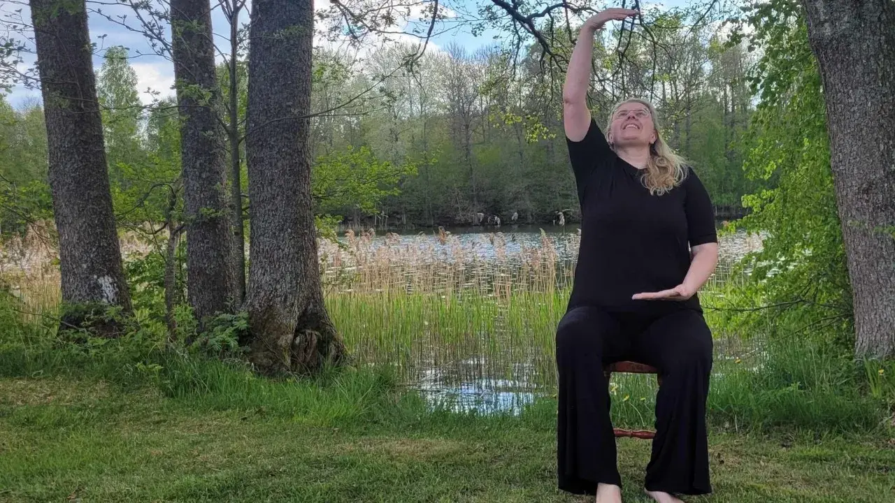 Kvinna i svart klädsel utför yoga på en stol vid en sjö. Naturen är grönskande med träd och vass.