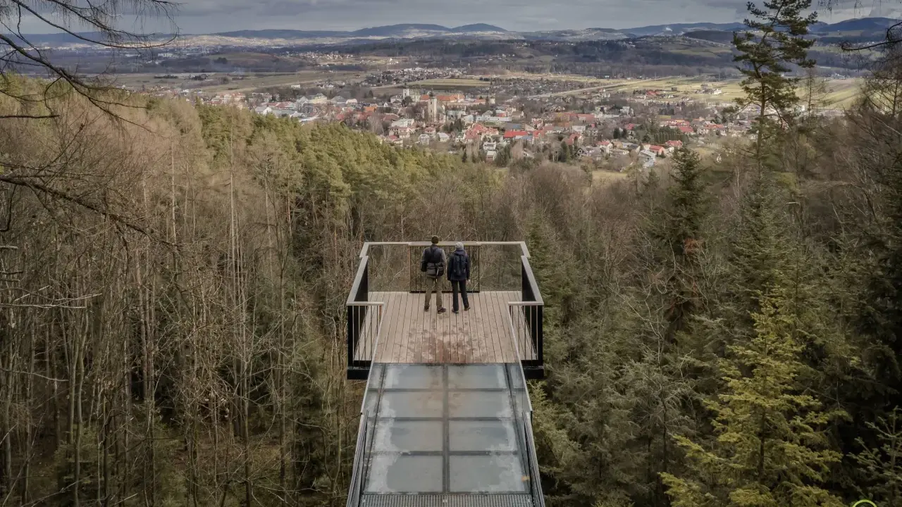 Dwie osoby stoją na szklanym tarasie widokowym, podziwiając panoramę Starego Sącza i okolic.