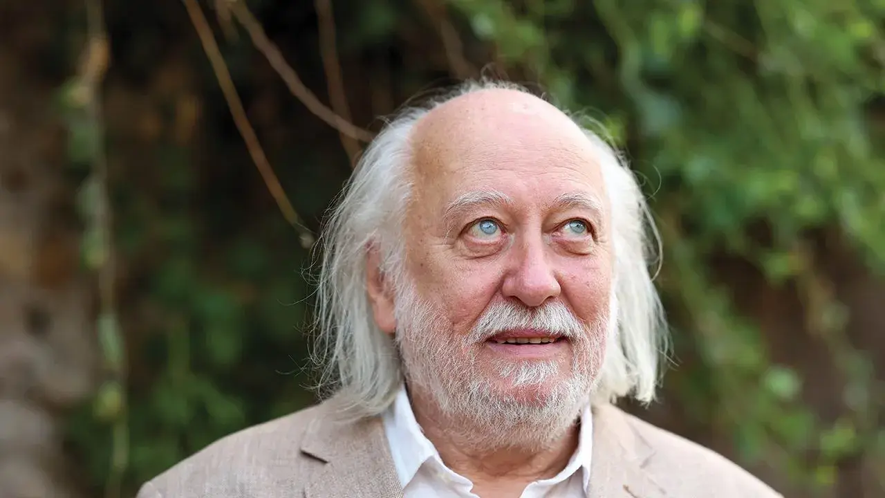 Ritratto di Laszlo Krasznahorkai, scrittore ungherese, con capelli bianchi e barba, sguardo pensieroso rivolto verso l'alto.