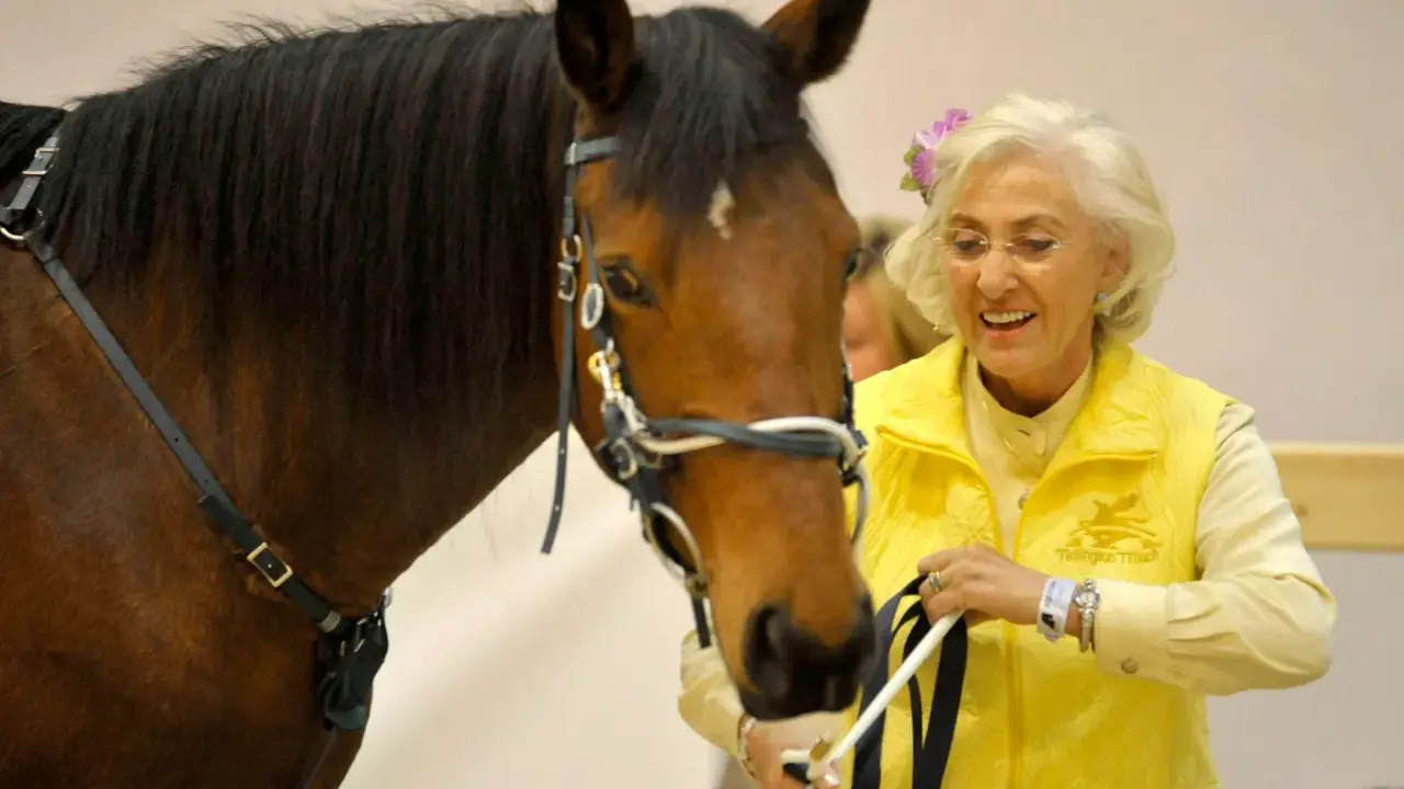 Linda Tellington mit einem braunen Pferd, sie trägt eine gelbe Weste und eine Blume im Haar.